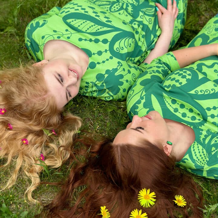 Kuvassa kaksi tyttöä makoilevat nurmikolla kukkia hiuksissaan ja päällään vihreät Krumeluuri kuosiset vaatteet